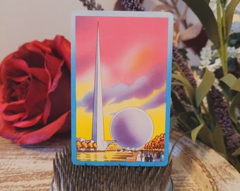 May include: A vintage postcard featuring a colorful illustration of a tall, white spire and a large, spherical structure against a sunset sky. The card has a blue border and depicts a landscape with trees and figures.