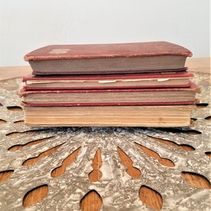 May include: A stack of four vintage hardcover books with red and brown covers. The books are sitting on a decorative metal surface with a circular pattern.