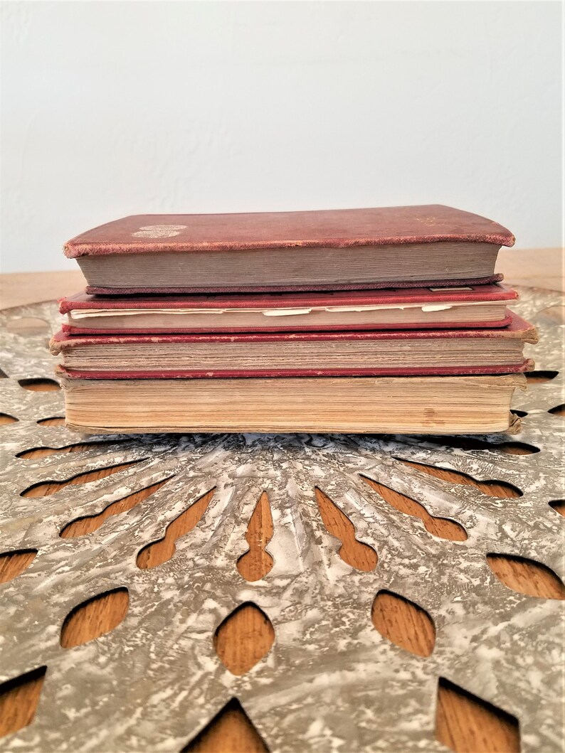 May include: A stack of four vintage hardcover books with red and brown covers. The books are sitting on a decorative metal surface with a circular pattern.