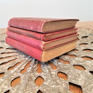 May include: A stack of four vintage hardcover books with red covers. The books have faded lettering on the spines and are slightly worn. The books are sitting on a decorative metal surface.