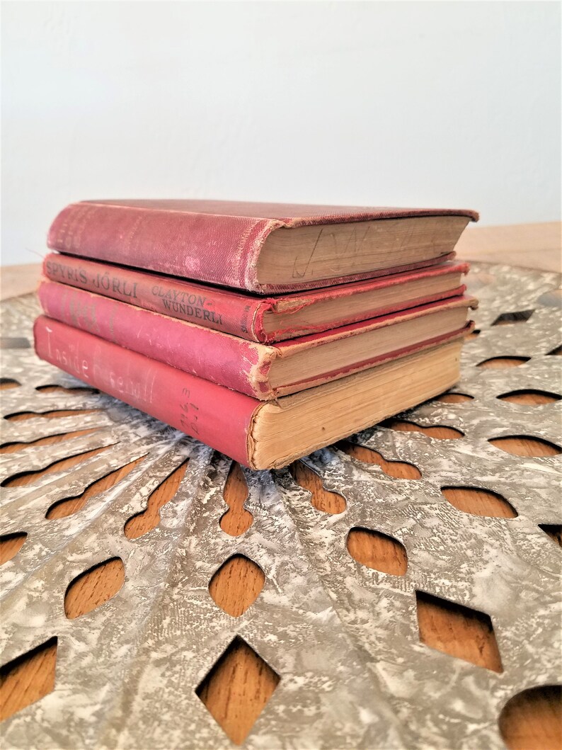 May include: A stack of four vintage hardcover books with red covers. The books have faded lettering on the spines and are slightly worn. The books are sitting on a decorative metal surface.