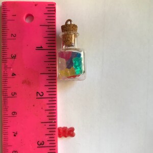 May include: A small glass jar with a cork stopper filled with colorful gummy bears. The jar is about 1 inch tall and is hanging from a silver chain. A single gummy bear is shown next to a ruler for scale.