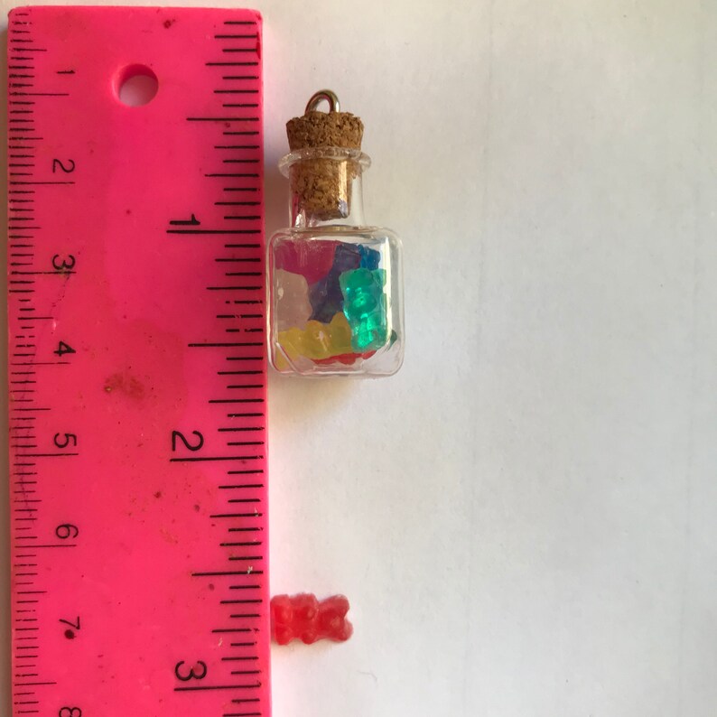 May include: A small glass jar with a cork stopper filled with colorful gummy bears. The jar is about 1 inch tall and is hanging from a silver chain. A single gummy bear is shown next to a ruler for scale.