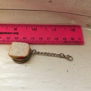 May include: A silver keychain with a miniature sandwich charm. The sandwich is made of white bread, lettuce, tomato, and a pickle. The keychain is about 2.5 inches long.
