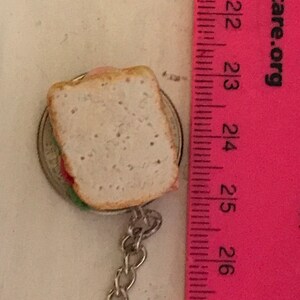 May include: A miniature sandwich keychain with a silver chain. The sandwich is made of two slices of white bread with a pink filling and green lettuce. The sandwich is about 1 inch in length and is attached to a silver chain.