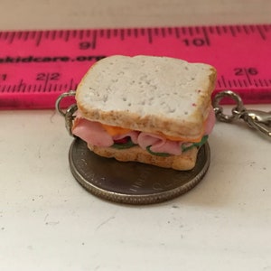 May include: A miniature clay sandwich keychain with a ham and cheese filling. The keychain is attached to a silver metal ring and is sitting on top of a US quarter.