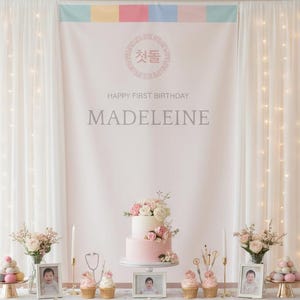 May include: A decorated table with a two-tiered pink and white cake, cupcakes, and framed photos. The backdrop features a banner with text in Korean and English, and the name "Madeleine". White curtains and string lights frame the scene.