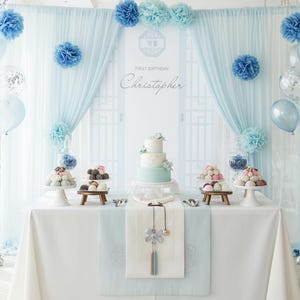 May include: A first birthday party setup with a light blue and white theme. The table is decorated with a cake, desserts, and a banner that reads "First Birthday Christopher". Balloons and paper decorations add to the festive atmosphere.