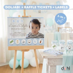 Puede incluir: Un juego de fiesta Doljabi en azul claro y blanco con boletos de rifa y etiquetas. El juego incluye un letrero con una foto de bebé y texto, frascos y una pila de boletos. El letrero está en un caballete de madera.