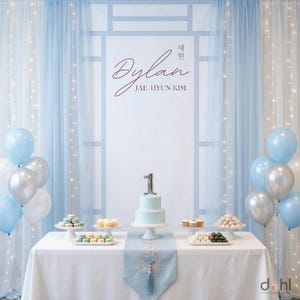 May include: A light blue and white party setup with a table, cake, and balloons. The table is set with desserts and a cake with a number one on top. The backdrop features sheer curtains and the name "Dylan" in cursive.