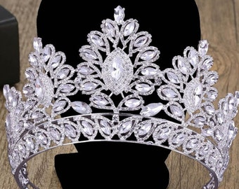 May include: A silver-toned crown with clear, sparkling crystals. The crown features a central teardrop-shaped crystal and is displayed on a black stand with the text "BEPHORA". The design is intricate and elegant, suitable for special occasions.