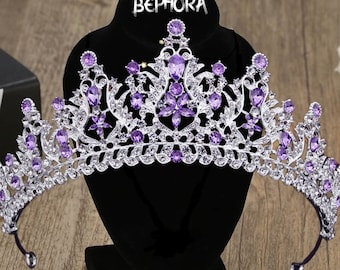 May include: A silver and purple tiara with a large, ornate design. The tiara is decorated with rhinestones and purple gems. The tiara is on a black stand with the word "BEPHORA" printed on it.