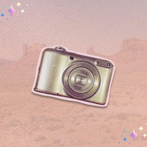 May include: A gold-colored digital camera with a prominent lens and the text "5X Wide Optical Zoom". The camera is set against a pink and brown background with a desert landscape.
