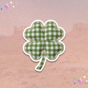 May include: A green and white gingham four-leaf clover sticker with a white border. The clover is made of fabric and has a stitched edge. The background is a gradient of pink and peach.