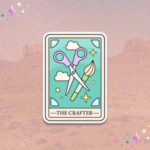 May include: A sticker featuring a teal tarot card design with scissors, a paintbrush, clouds, and stars. The text "THE CRAFTER" is at the bottom. The sticker has a white border and is set against a pink and brown background.