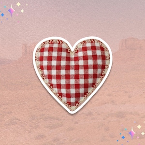 May include: A heart-shaped sticker with a red and white gingham pattern. The sticker has a white border and a fringed edge. The background is a soft pink with a desert landscape.