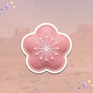 May include: A pink felt flower-shaped applique with white stitching and a white snowflake design in the center. The applique has a white border and is set against a soft, textured background.