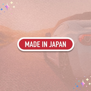 May include: A red and white oval sticker with the text "MADE IN JAPAN" is displayed against a blurred background featuring a car and a road. The sticker has a white border and is set against a gradient of pink and orange hues.