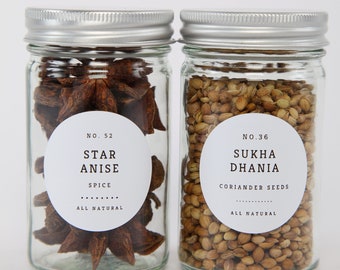 May include: Two glass jars with silver lids filled with spices. The jar on the left contains star anise and has a label that reads "No. 52 Star Anise Spice All Natural". The jar on the right contains coriander seeds and has a label that reads "No. 36 Sukha Dhani Coriander Seeds All Natural".