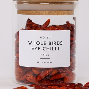 Op de afbeelding: Een glazen pot met een bamboe deksel gevuld met gedroogde rode bird's eye chili pepers. De pot heeft een wit etiket met de tekst "No. 59 Whole Birds Eye Chilli Spice All Natural".