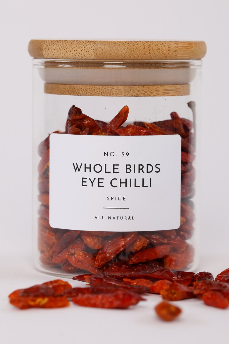 Op de afbeelding: Een glazen pot met een bamboe deksel gevuld met gedroogde rode bird's eye chili pepers. De pot heeft een wit etiket met de tekst "No. 59 Whole Birds Eye Chilli Spice All Natural".