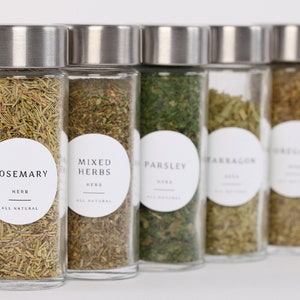 Peut inclure: Six bocaux en verre avec des couvercles argentés remplis d'herbes séchées. Les bocaux sont étiquetés avec des étiquettes circulaires blanches qui indiquent "Romarin", "Herbes mélangées", "Persil", "Estragon" et "Origan".