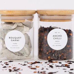 Op de afbeelding: Twee glazen potten met bamboe deksels gevuld met gedroogde kruiden en specerijen. De pot links bevat gedroogde kruiden met het label "Bouquet Garni Kruid Alles Natuurlijk". De pot rechts bevat gedroogde ancho chili vlokken met het label "Nr. 01 Ancho Chili Specerij Alles Natuurlijk".