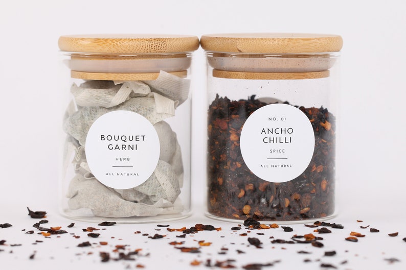Op de afbeelding: Twee glazen potten met bamboe deksels gevuld met gedroogde kruiden en specerijen. De pot links bevat gedroogde kruiden met het label "Bouquet Garni Kruid Alles Natuurlijk". De pot rechts bevat gedroogde ancho chili vlokken met het label "Nr. 01 Ancho Chili Specerij Alles Natuurlijk".