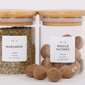 Op de afbeelding: Twee glazen potten met bamboe deksels gevuld met gedroogde kruiden en hele nootmuskaat. De potten hebben witte etiketten met zwarte tekst die "NO. 36 MARJORAM HERB ALL NATURAL" en "NO. 60 WHOLE NUTMEG SPICE ALL NATURAL" luidt.