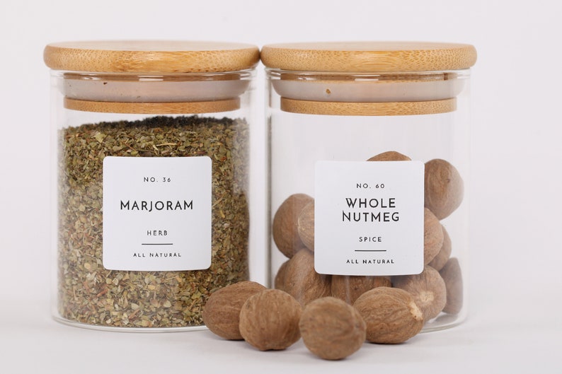 Op de afbeelding: Twee glazen potten met bamboe deksels gevuld met gedroogde kruiden en hele nootmuskaat. De potten hebben witte etiketten met zwarte tekst die "NO. 36 MARJORAM HERB ALL NATURAL" en "NO. 60 WHOLE NUTMEG SPICE ALL NATURAL" luidt.