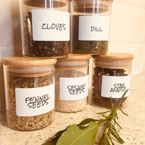 Puede incluir: Cinco tarros de vidrio con tapas de madera llenos de diferentes especias. Los tarros están etiquetados con etiquetas escritas a mano: Clavos, Eneldo, Semillas de hinojo, Semillas de sésamo y Anís estrellado.
