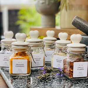 Op de afbeelding: Een set van zes glazen kruidenpotten met witte keramische deksels en hartvormige handvatten. Elke pot heeft een zwart-wit etiket met de naam van het kruid en het nummer van de pot. De kruiden zijn laurierblaadjes, gemalen kurkuma, mosterdzaad, korianderzaad, sesamzaad en chilivlokken.