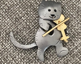 May include: A silver-colored cat-shaped brooch playing a golden violin. The cat has etched details, including whiskers and stripes. The violin has a detailed shape, and the bow is a thin, straight line. The brooch is set against a textured gray background.