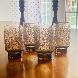 May include: Four amber-colored glass tumblers with a delicate gold swirl design. The glasses have a rounded bottom and a wide mouth.