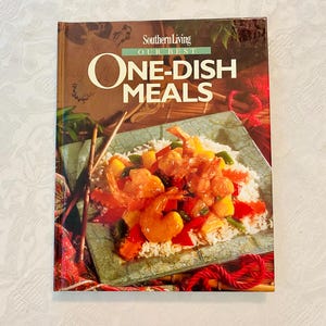 May include: A cookbook titled "Southern Living Our Best One-Dish Meals" features a vibrant image of a shrimp and vegetable dish on a bed of rice. The cover is predominantly brown with green and red accents, and the title is in large, bold letters.