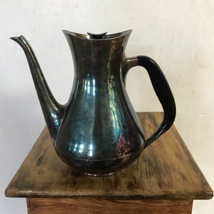 May include: A vintage silver-toned coffee pot with a long spout and a black handle. The pot has a curved body and a small black lid. It sits on a wooden table.