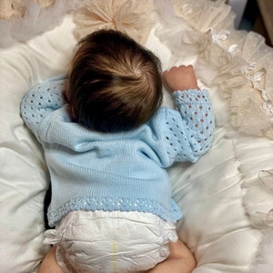 Könnte beinhalten: Eine Babypuppe, die einen hellblauen Strickpullover mit einer weißen Windel trägt. Die Puppe liegt auf dem Rücken auf einer weißen und beigefarbenen Stoffoberfläche.