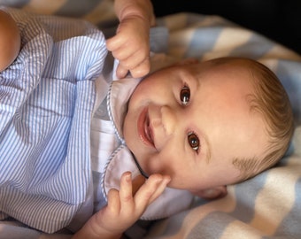 Puede incluir: Primer plano de una muñeca bebé con ojos marrones, una peluca marrón claro y un atuendo a rayas azules y blancas. La muñeca está tumbada sobre una manta a rayas azules y blancas.