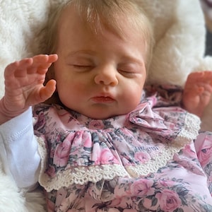 Könnte beinhalten: Eine schlafende Baby-Puppe, die ein rosa Blumenkleid mit weißer Spitzenborte trägt.