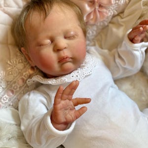 Könnte beinhalten: Eine lebensechte Babypuppe mit geschlossenen Augen, rosigen Wangen und hellbraunem Haar. Die Puppe trägt einen weißen Strampler mit Spitzenkragen und eine rosa Schleife. Sie liegt auf einer weiß-rosa gemusterten Decke.