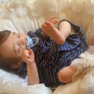Könnte beinhalten: Eine schlafende Babypuppe mit braunen Haaren, die ein blaues und weiß gemustertes Outfit und einen blauen Schnuller trägt. Die Puppe liegt auf einem weißen Spitzenstoff.