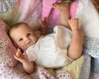 May include: A realistic baby doll with brown eyes and a sweet smile, wearing a white dress with a pink trim, lying on a colorful patchwork quilt.