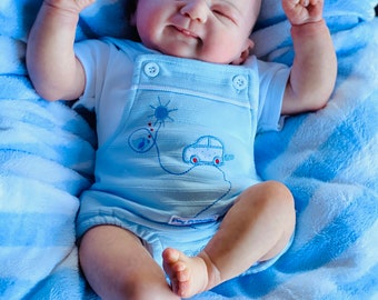 Puede incluir: Un muñeco bebé con un mono azul claro con un diseño de coche y una camiseta blanca. El muñeco está tumbado sobre una manta de rayas azules y blancas.