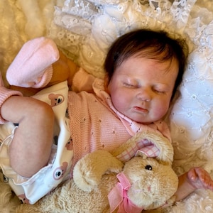 Op de afbeelding: Een realistische pasgeboren babypop met donker haar, slapend met gesloten ogen. De pop draagt een lichtroze gebreid topje, een wit luierbroekje met berenprint en lichtroze geribbelde sokjes. Het houdt een klein, beige pluche konijntje vast.
