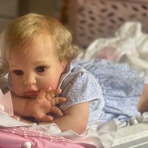 May include: A realistic baby doll with blonde hair and blue eyes, wearing a light blue and white floral dress, lying on a pink and white patterned blanket.
