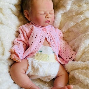 May include: A lifelike baby doll with closed eyes, wearing a light pink knitted cardigan and a diaper. The doll is lying on a soft, white, textured blanket. The cardigan has a delicate, openwork design.