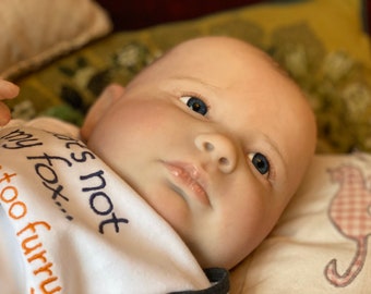 Puede incluir: Primer plano de una muñeca bebé con ojos azules y una tez rosa claro. La muñeca lleva un babero blanco con el texto "No es mi zorro...  somos demasiado peludos".