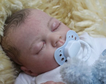 Puede incluir: Un muñeco bebé durmiendo con los ojos cerrados y un chupete azul. El muñeco lleva un pelele blanco y está tumbado en una manta blanca esponjosa.