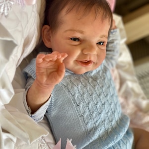 Può includere: Una bambola di bambino sorridente con i capelli castani che indossa un maglione di lana blu chiaro con un motivo a treccia.
