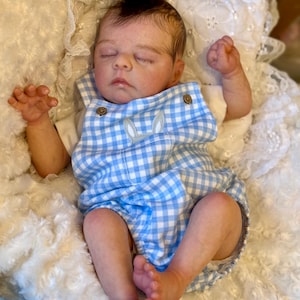 Può includere: Una bambola realistica di neonato con capelli scuri e occhi chiusi, che indossa una tutina a quadretti vichy azzurri e bianchi con due piccoli bottoni marroni e un dettaglio ricamato di orecchie di coniglio sul petto.